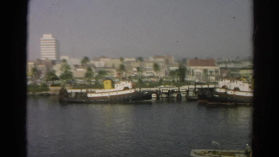 1966 8mm view across a California waterfront toward docked boats, palm-lined buildings, and a white high-rise under a hazy sky.