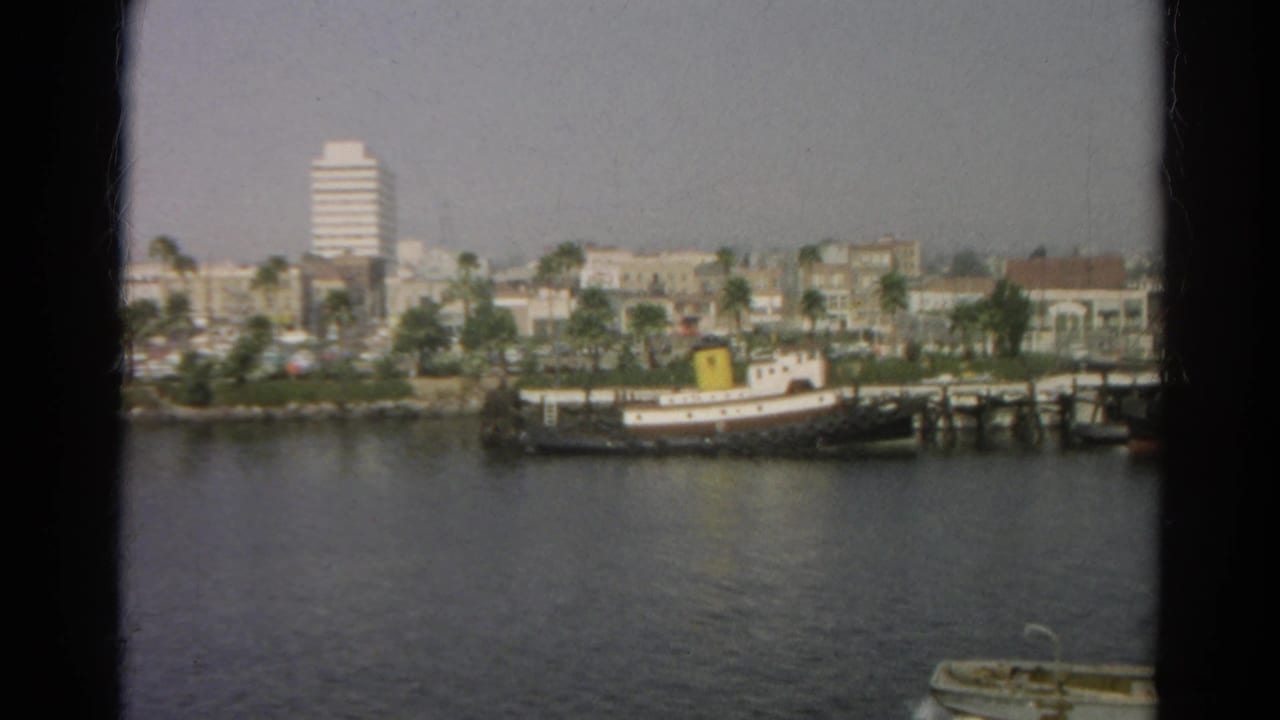 City shoreline across water with a docked tugboat, pier, palms, and a white high-rise in the background.