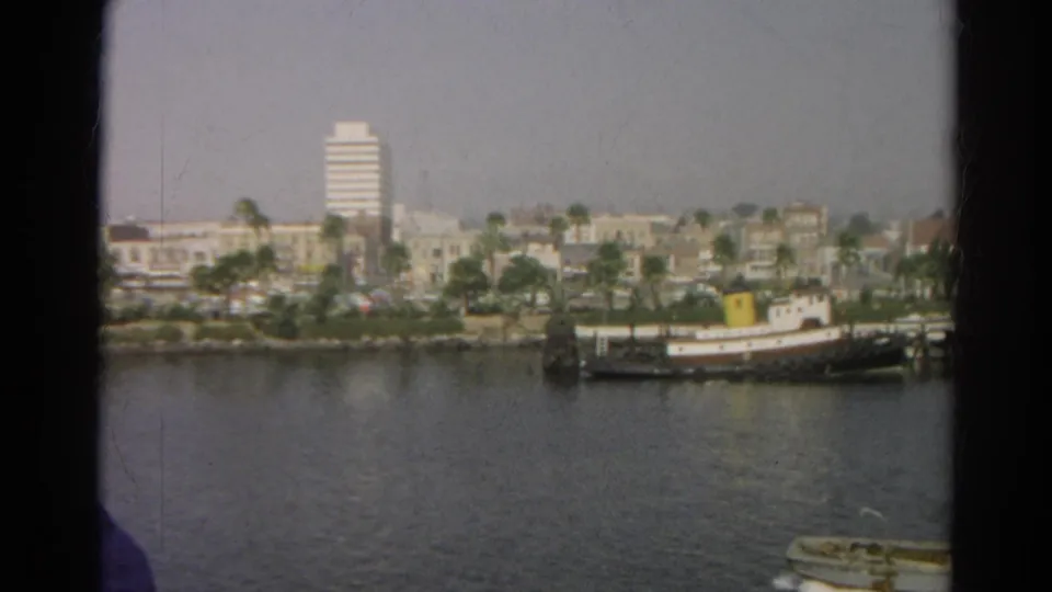 1966 home-movie frame from a boat ride shows water, a docked workboat, palms, low-rise shoreline buildings, and a white high-rise.