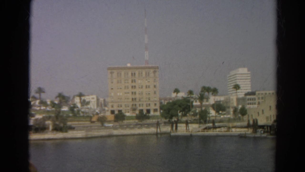 Hazy 1966 Silver Lake waterfront view with a central multi-story building, mast, palm-lined shore, and a distant modern high-rise.