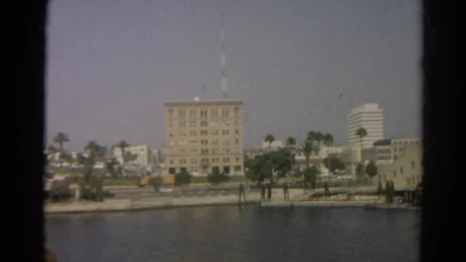 Hazy 1966 Silver Lake waterfront view with a central multi-story building, mast, palm-lined shore, and a distant modern high-rise.