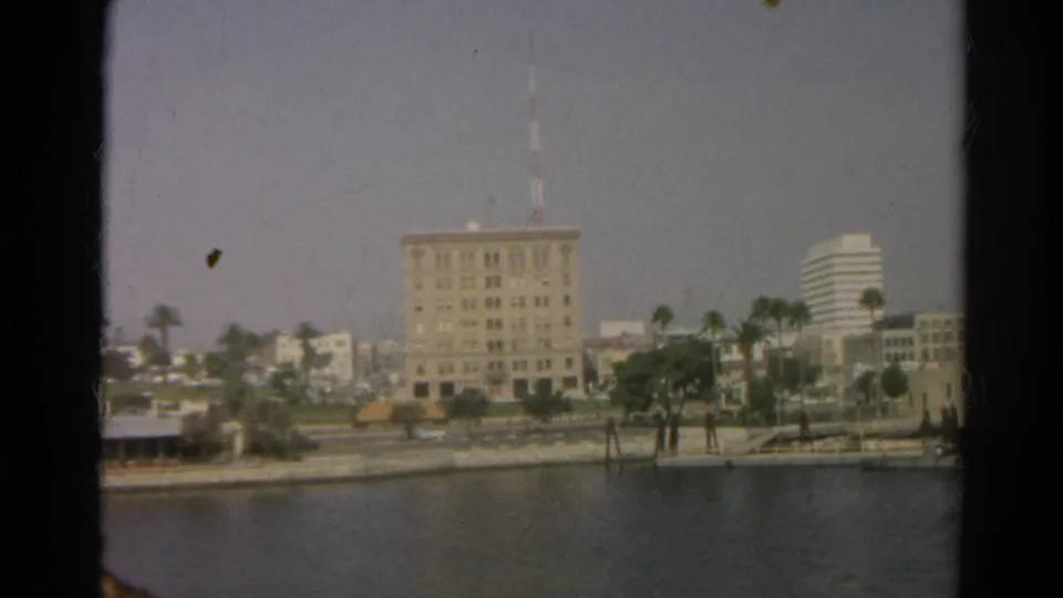 1966 Silver Lake shoreline seen across the water, with palm trees, a prominent central building, a mast, and distant city buildings.