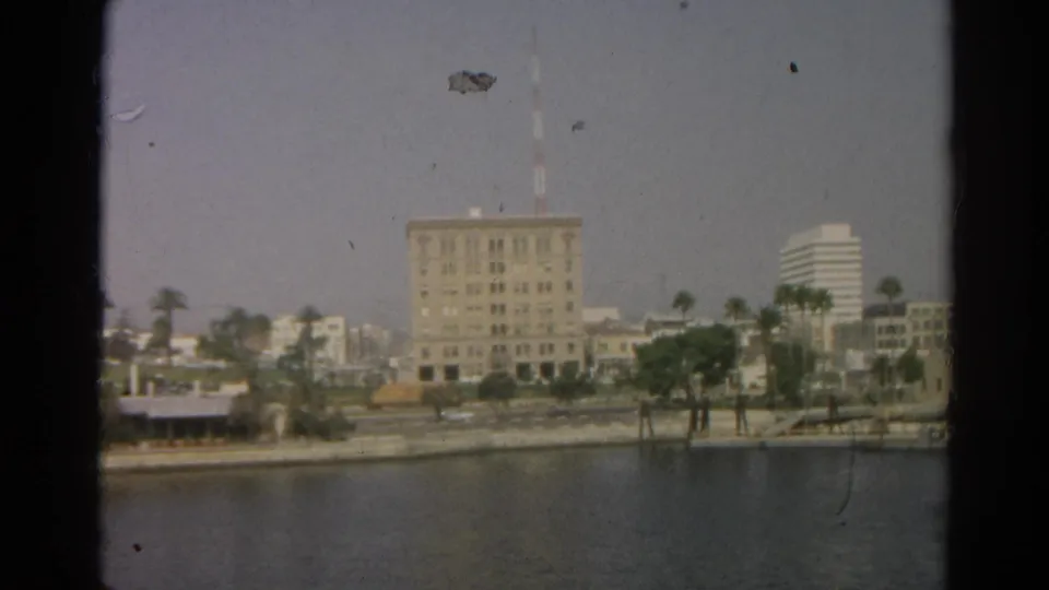 Hazy 1966 Silver Lake waterfront frame with a central towered building, palm trees, water in the foreground, and visible 8mm dust marks.