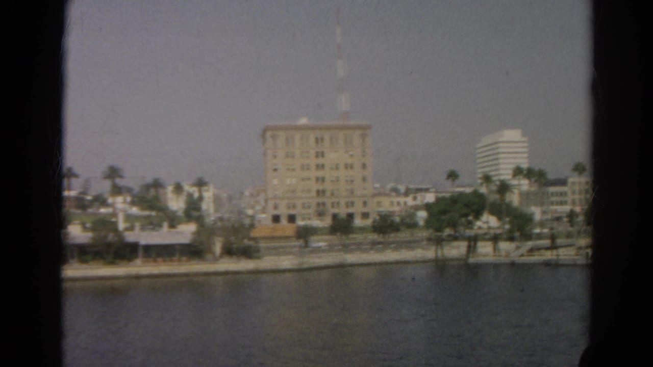 Boat-view frame of the Silver Lake shoreline with a large building, palms, and low waterfront structures in soft 8mm color.