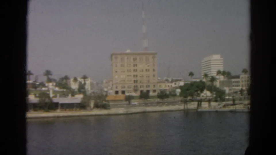 Boat-view frame of the Silver Lake shoreline with a large building, palms, and low waterfront structures in soft 8mm color.