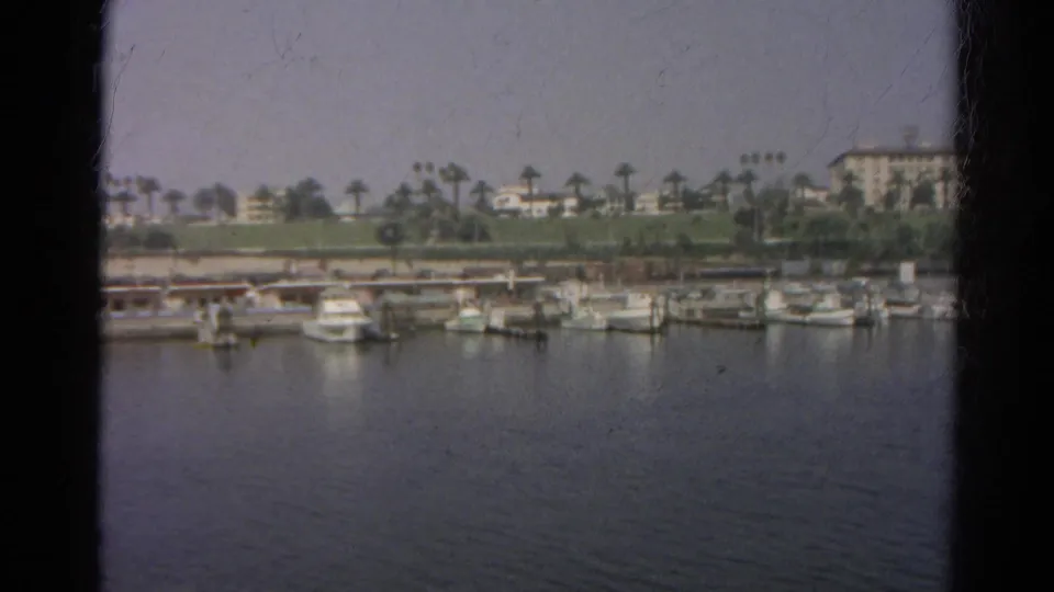 1966 California waterfront frame showing a marina with docked small boats, palms, hillside lawns, and buildings in the background.