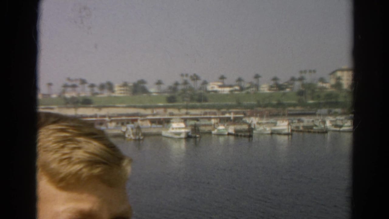 1966 8mm California harbor view from a boat window, with small boats docked across calm water and a partial passenger head in the foreground.
