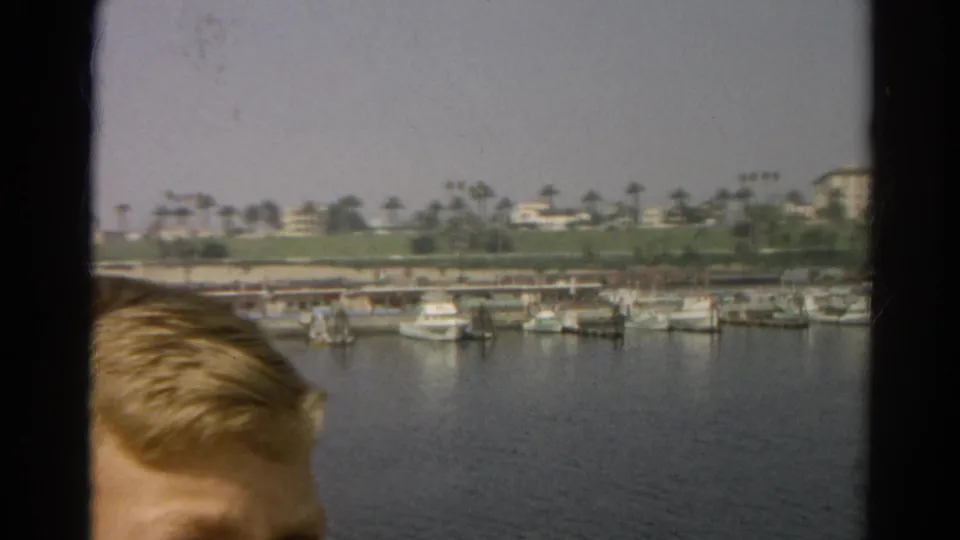 1966 8mm California harbor view from a boat window, with small boats docked across calm water and a partial passenger head in the foreground.