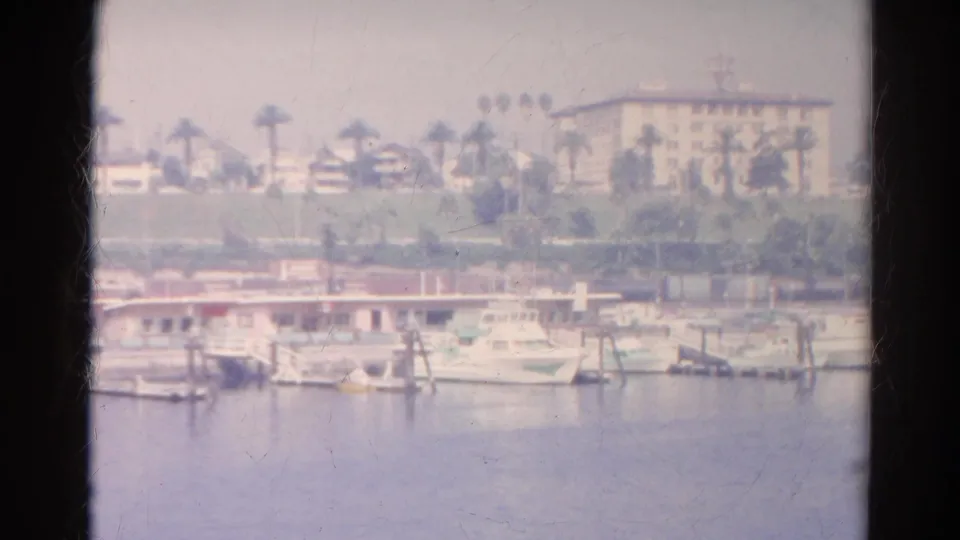 Faded 1966 8mm harbor frame showing small boats, marina structures, palms, and waterfront buildings beyond calm water.