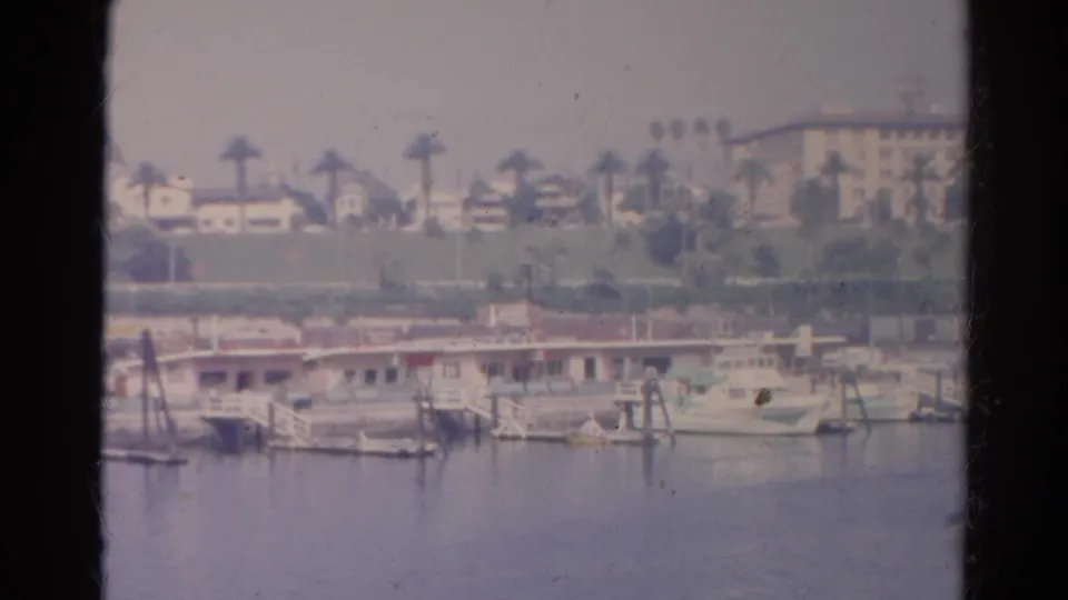 1966 8mm California marina view from a boat, with docked craft, palm trees, hillside buildings, and a dark window divider.