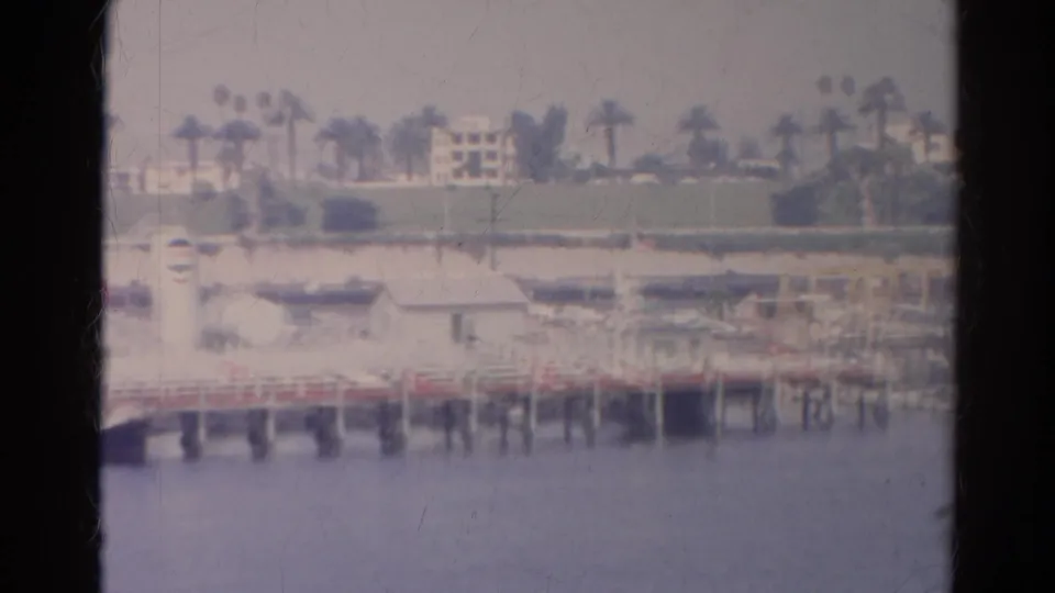 Bright 8mm view of Silver Lake waterfront, with a pier, small buildings, palms, and homes across the water.