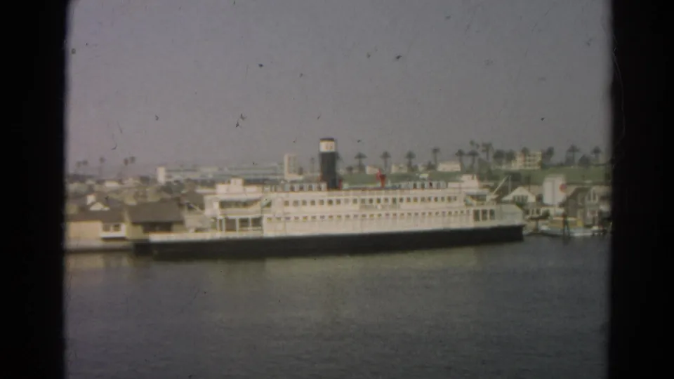 Soft 1966 8mm view of a large docked passenger ship on harbor water, with a hazy row of palm trees and buildings behind it.