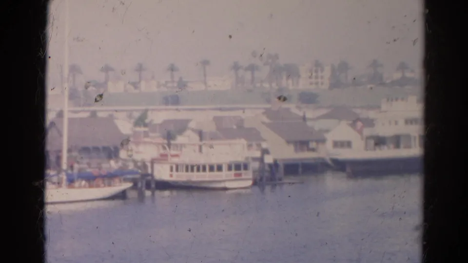 1966 home-movie frame showing small boats, dock structures, palms, and waterfront buildings in the listed California location.