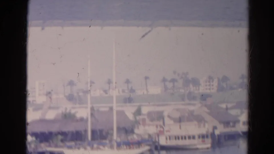 Faded 1966 8mm frame of a hazy waterfront view with boat masts, a docked vessel, palms, and low buildings.
