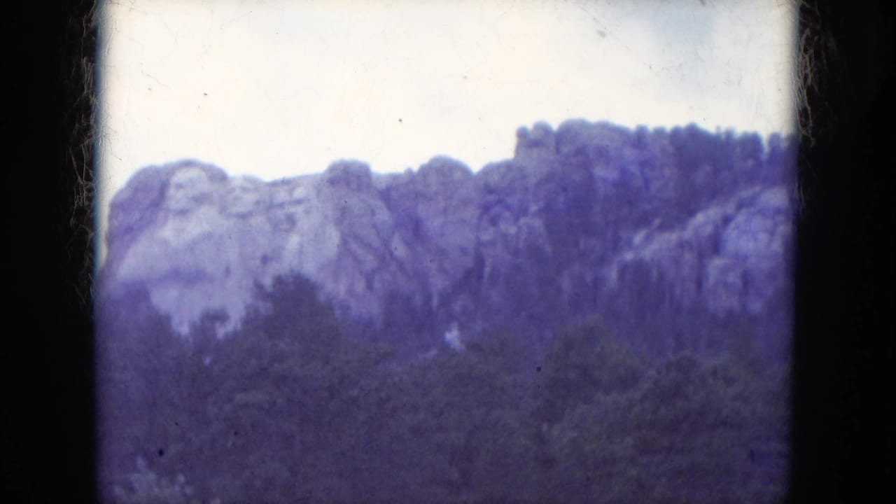 1966 8mm view of Mount Rushmore National Memorial above a dark tree line with visible film gate edges and blue-purple color cast.