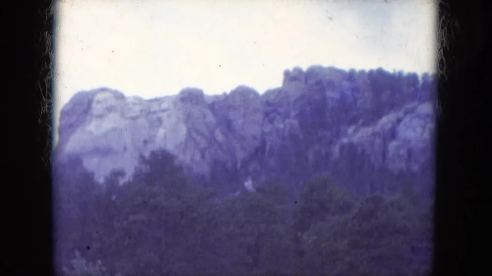 1966 8mm view of Mount Rushmore National Memorial above a dark tree line with visible film gate edges and blue-purple color cast.