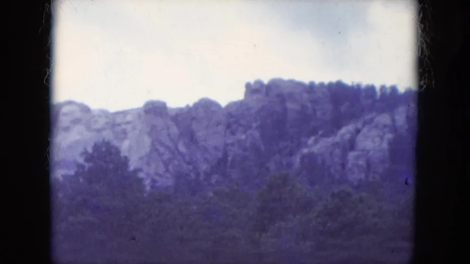 1966 home-movie frame showing the Mount Rushmore carvings across a rocky ridge, with trees below and dark 8mm frame borders.