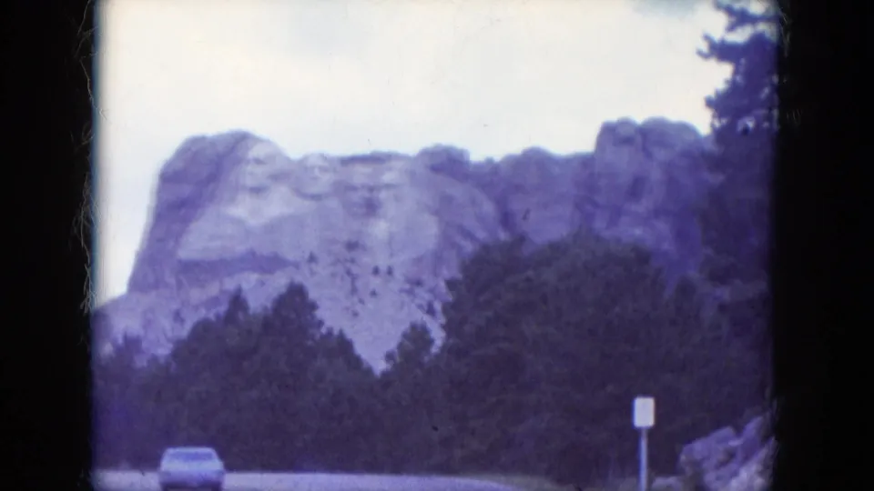 1966 8mm roadside view of Mount Rushmore, with a small car and sign below the tree line adding scale to the monument.