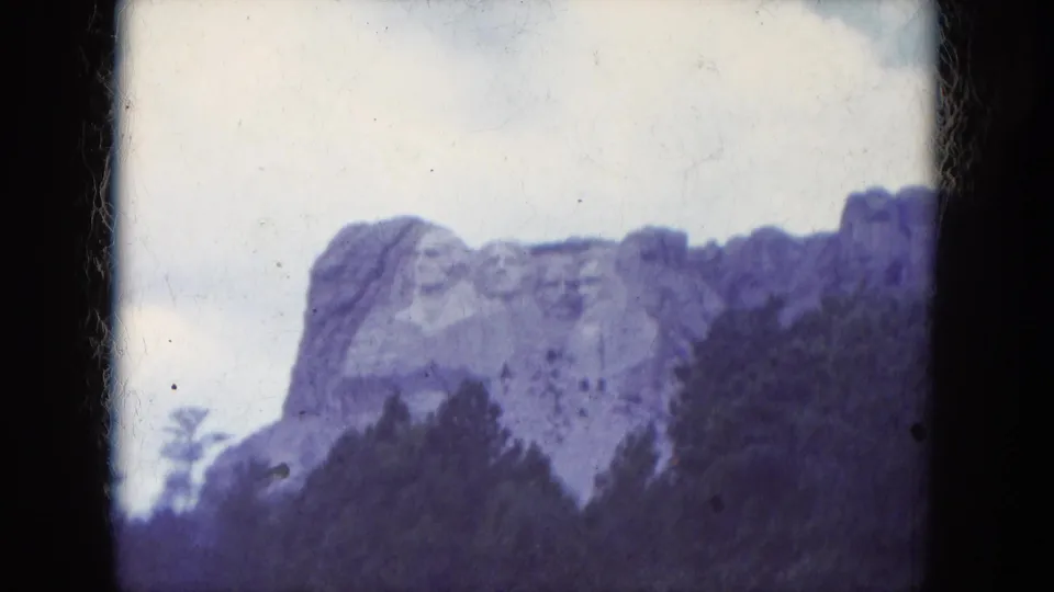 1966 8mm frame of Mount Rushmore seen through trees, with pale sky, visible dust marks, and dark film borders.