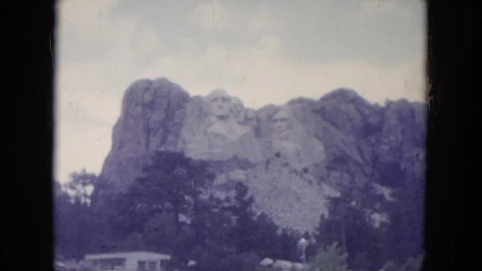 1966 visitor view of Mount Rushmore, showing the carved heads above trees with a small building and parked vehicles near the base.