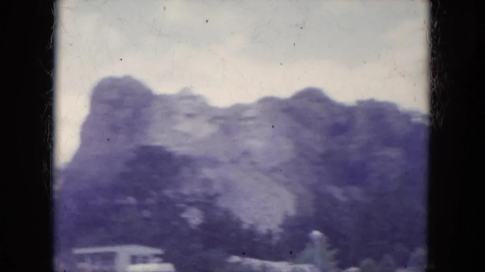Soft, purple-tinted 8mm frame of the Mount Rushmore carving, with trees crossing the lower foreground and cloudy sky above.