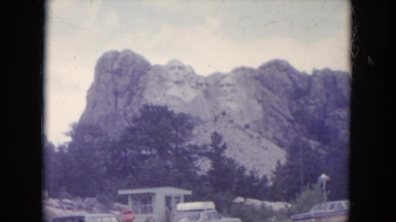 1966 8mm view of Mount Rushmore in South Dakota, framed with trees, cars, and a small building below the carved faces.