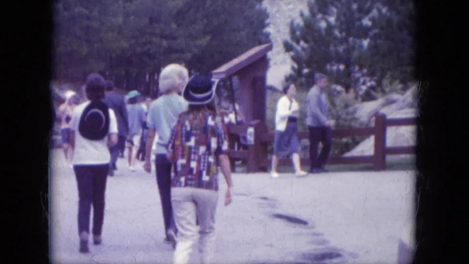 Tourists walk away from the camera at Mount Rushmore, passing a small structure, railing, trees, and other visitors.