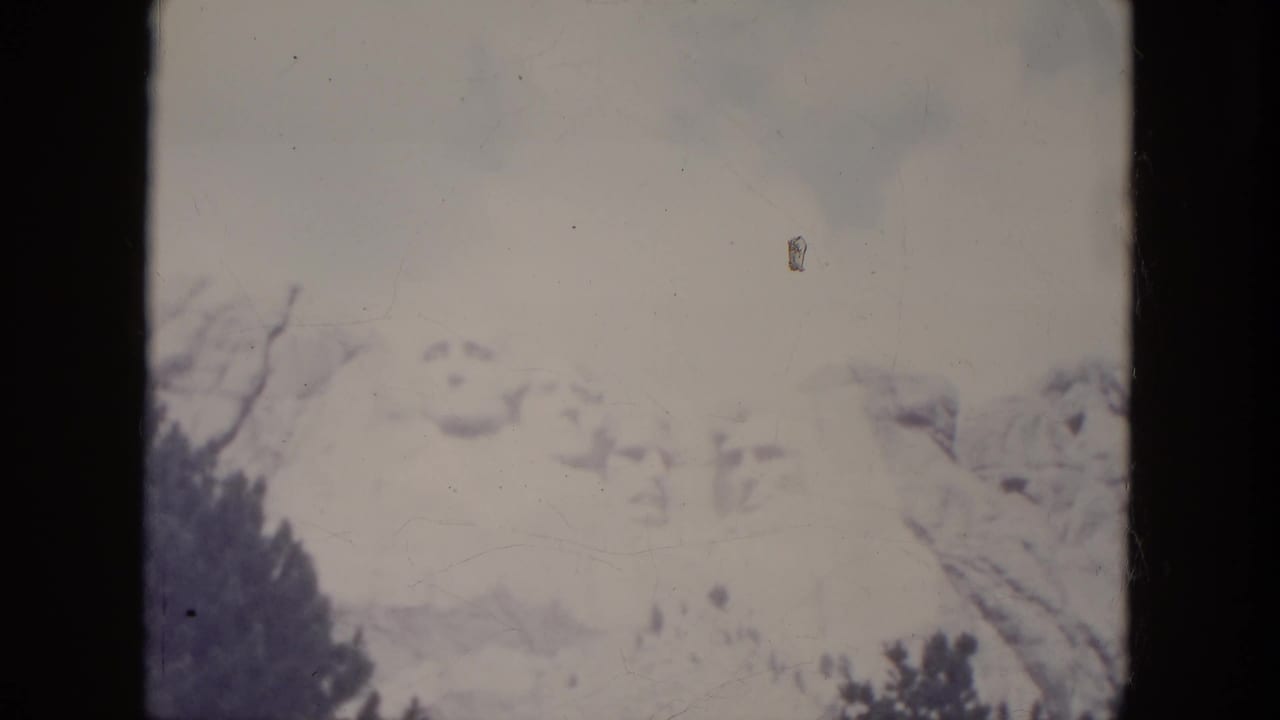 1966 8mm view of Mount Rushmore in South Dakota, with pale sky, soft presidential carvings, and dark trees below.