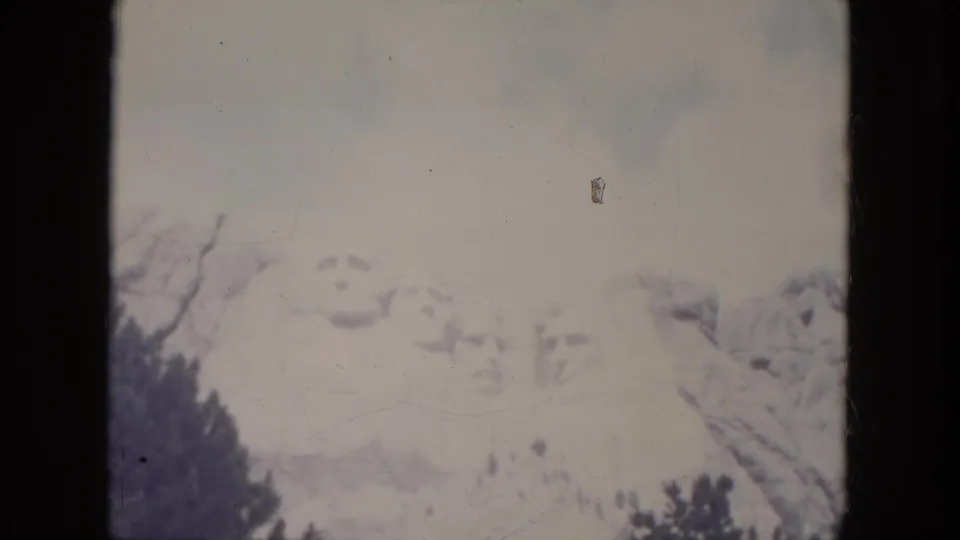 1966 8mm view of Mount Rushmore in South Dakota, with pale sky, soft presidential carvings, and dark trees below.