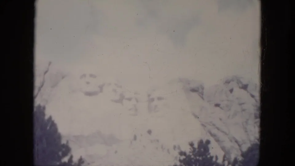 1966 South Dakota home-movie frame showing Mount Rushmore's carved faces under a pale sky with trees at the bottom.