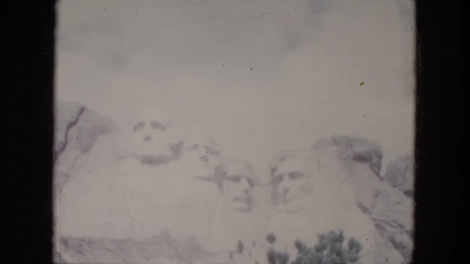 Vintage 8mm frame from 1966 showing Mount Rushmore partly low in the composition, with cloudy sky and trees in South Dakota.