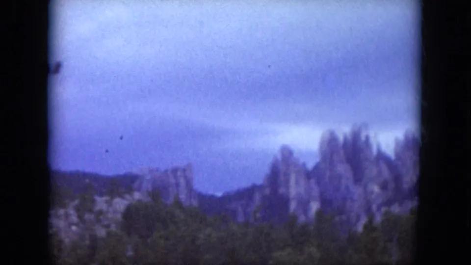 1966 South Dakota 8mm scenic frame showing rocky formations beyond trees under a cloudy sky.