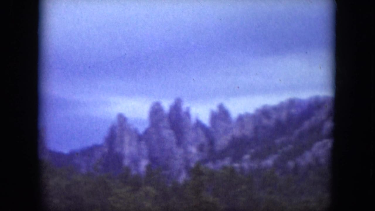 1966 South Dakota 8mm frame showing hazy rock formations beyond evergreen forest beneath an overcast sky.