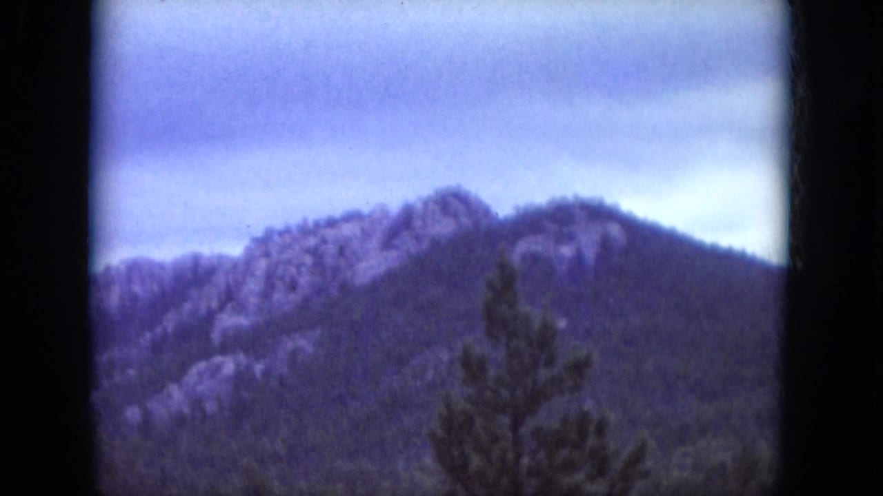 Tree-lined mountain ridge with rocky outcrops below a pale cloudy sky.