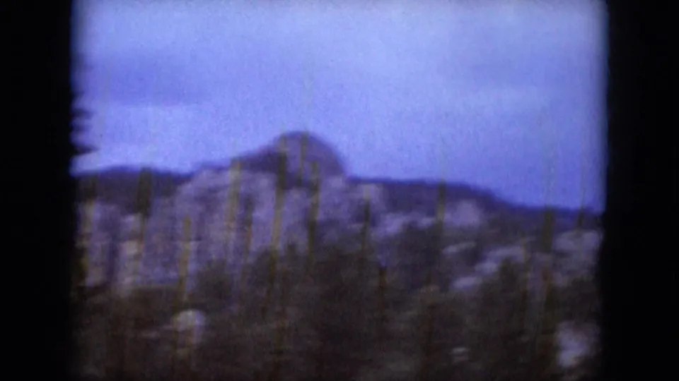 1966 South Dakota frame showing a rocky hillside, scattered slender trees, and darker forest below a cloudy sky.