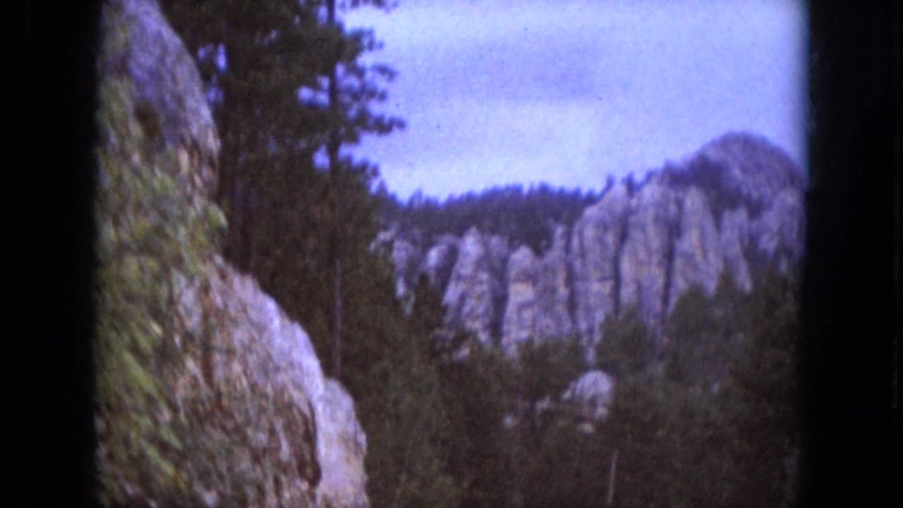 1966 South Dakota 8mm frame showing rugged rock formations, evergreens, and a pale sky with purple color shift.