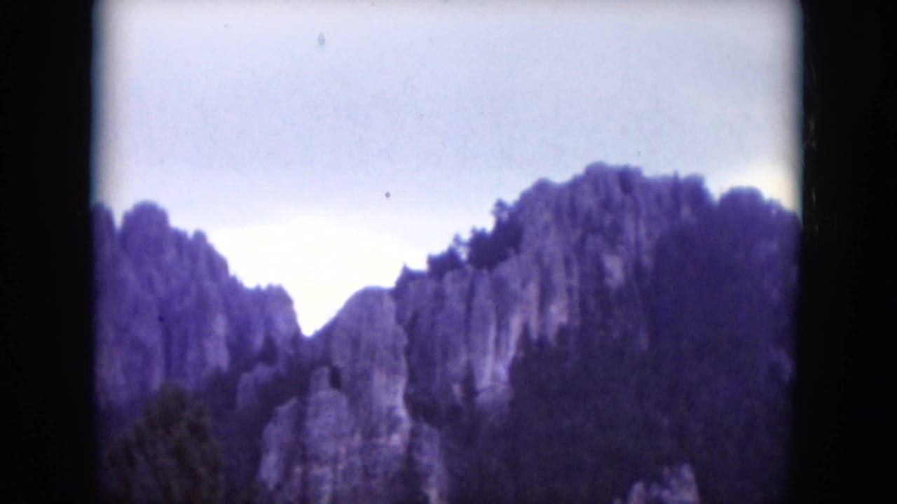 Distant rock spires and wooded slopes under a pale sky within an 8mm frame.