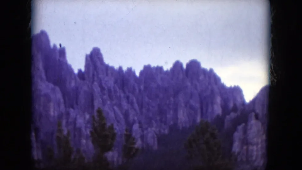 1966 South Dakota 8mm frame showing a jagged rock ridge above evergreens, softened by film grain and age shift.