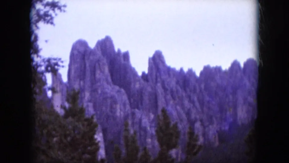 1966 South Dakota home-movie frame with tall rock formations, evergreen tops, and visible 8mm frame edges.