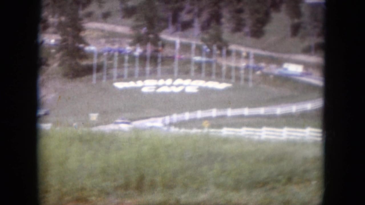 Grassy hillside with white Rushmore Cave sign, fencing, road, and cars.