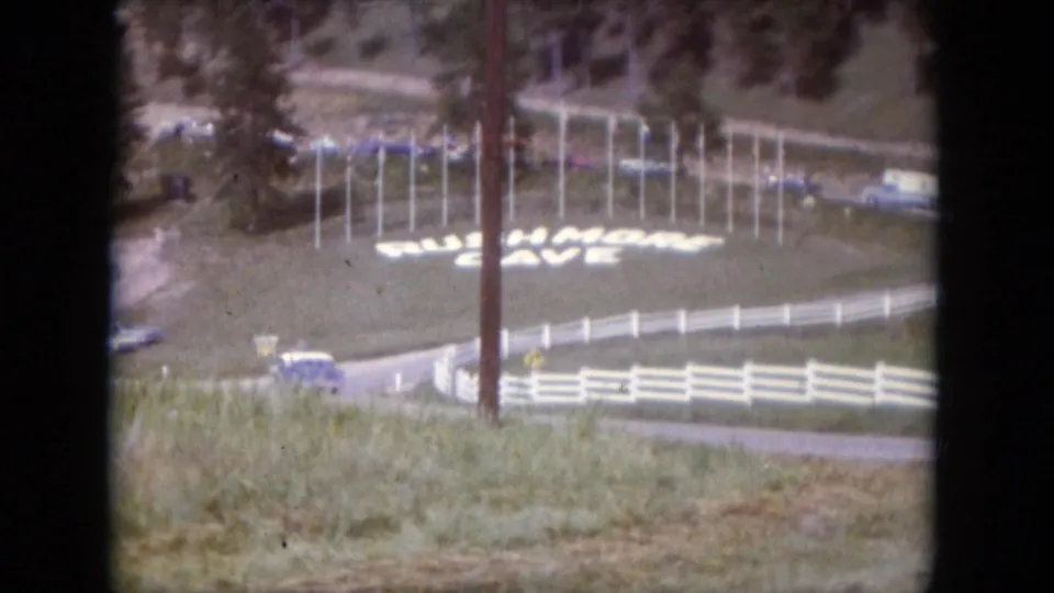 Visitor-shot 1966 roadside frame showing the Rushmore Cave sign, white fencing, classic cars, and a utility pole.