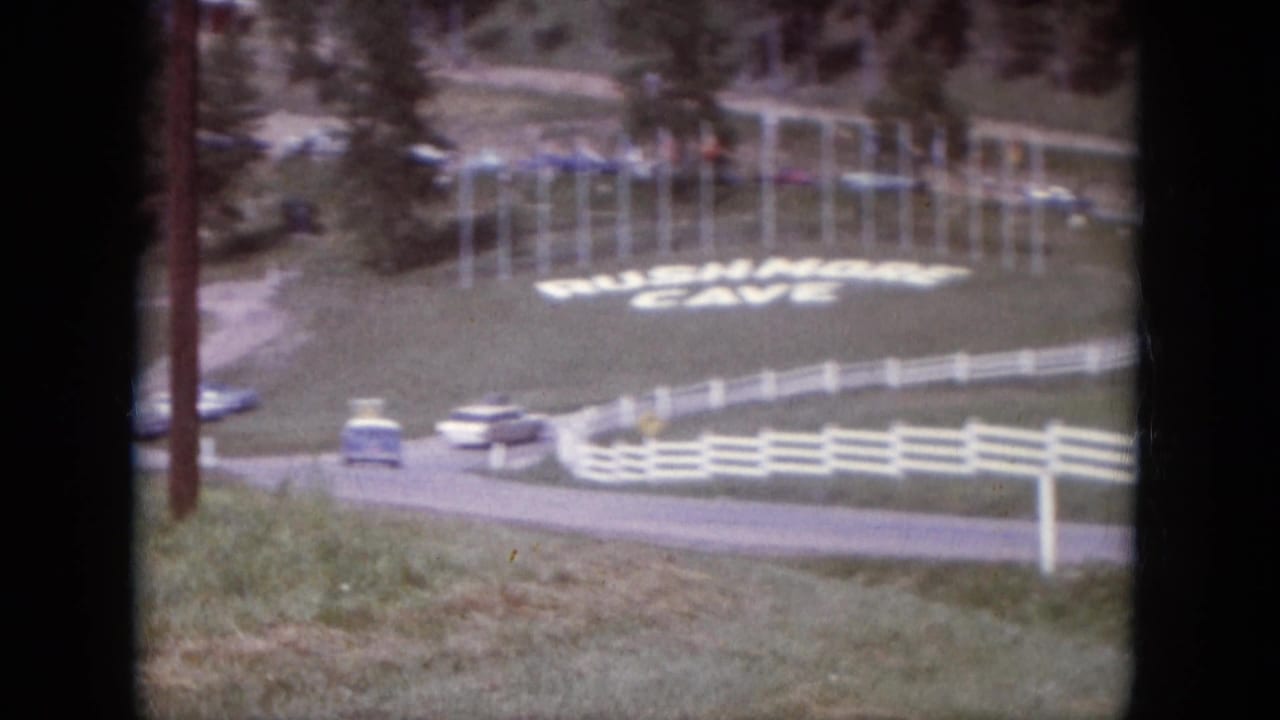 Curving road near white fences with cars and a Rushmore Cave sign on grass.