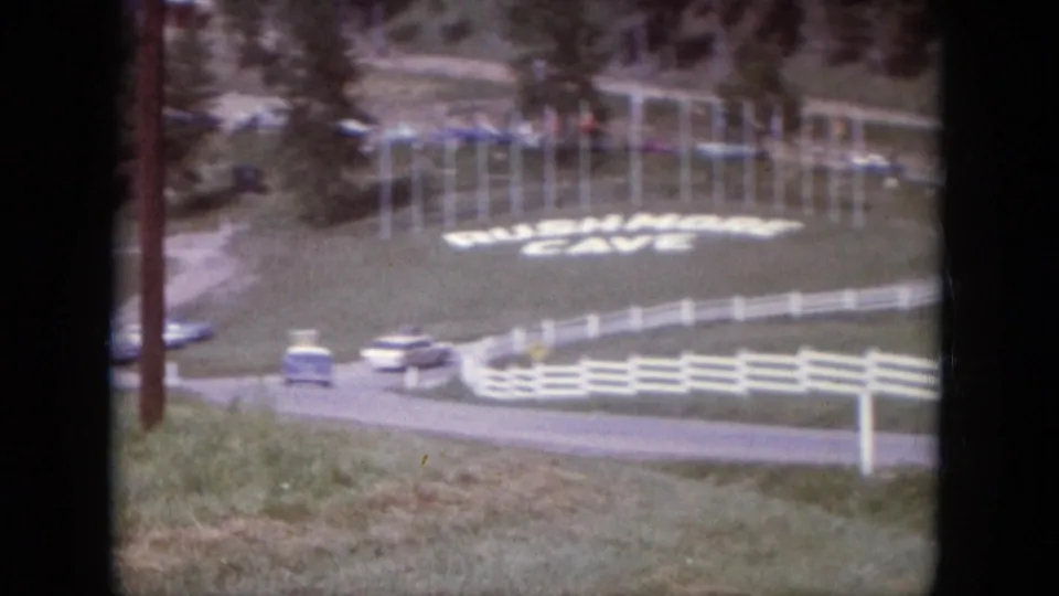 1966 South Dakota 8mm frame showing a road, white fencing, cars, trees, and a large Rushmore Cave sign on a grassy slope.