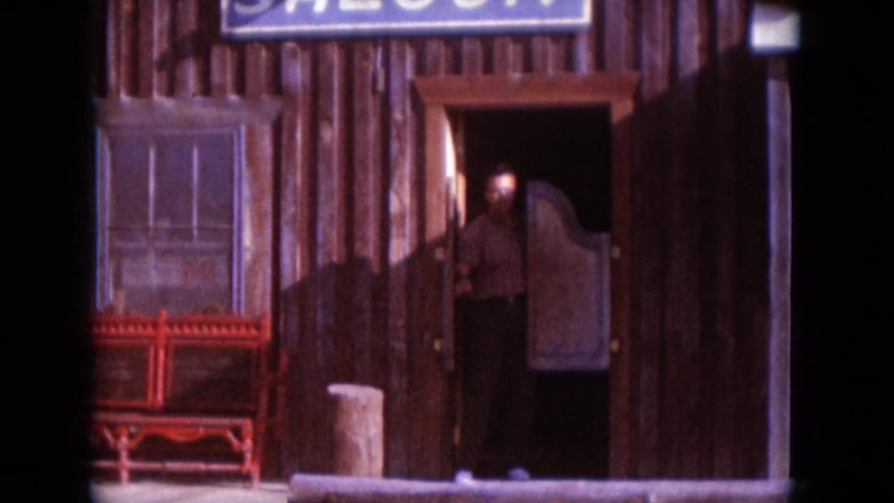 Adult man emerging from a rustic wooden saloon doorway in a 1966 South Dakota home-movie frame, with signage above and bench nearby.