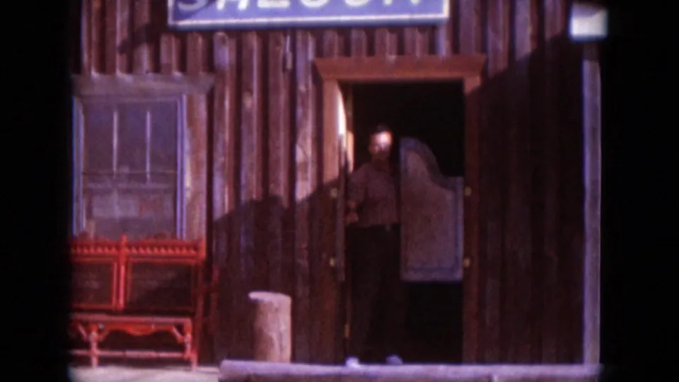 Adult man emerging from a rustic wooden saloon doorway in a 1966 South Dakota home-movie frame, with signage above and bench nearby.
