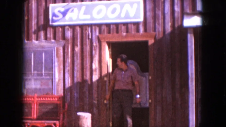 Adult man in a striped shirt stepping from a wooden saloon doorway, with a large saloon sign and red bench visible.