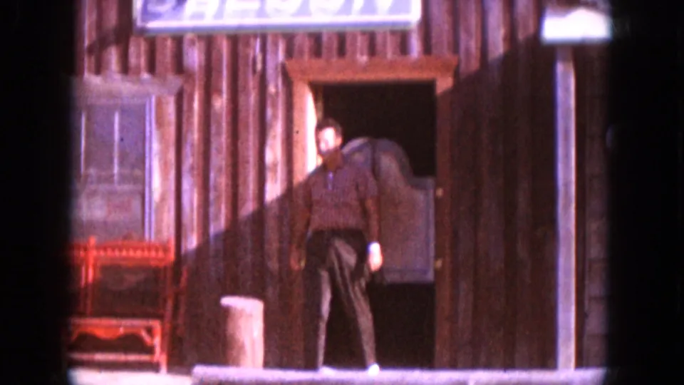 1966 South Dakota home-movie frame of an adult man walking from a rustic saloon entrance beneath a prominent saloon sign.