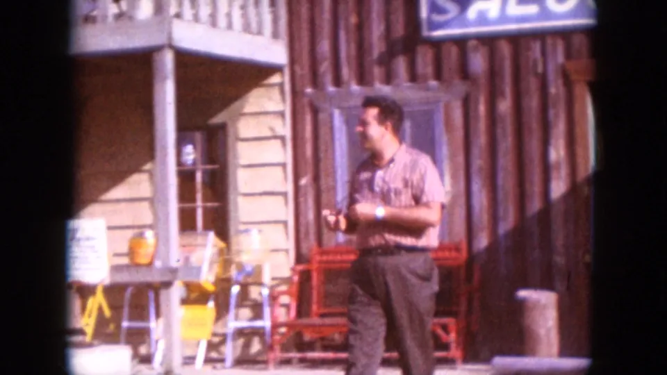 The man continues past the saloon facade, with the red bench, porch details, and adjacent light-colored building visible.