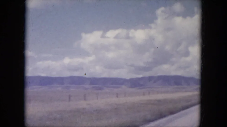 1960s 8mm view across South Dakota plains with fence posts, low hills, heavy clouds, and a road edge in the foreground.