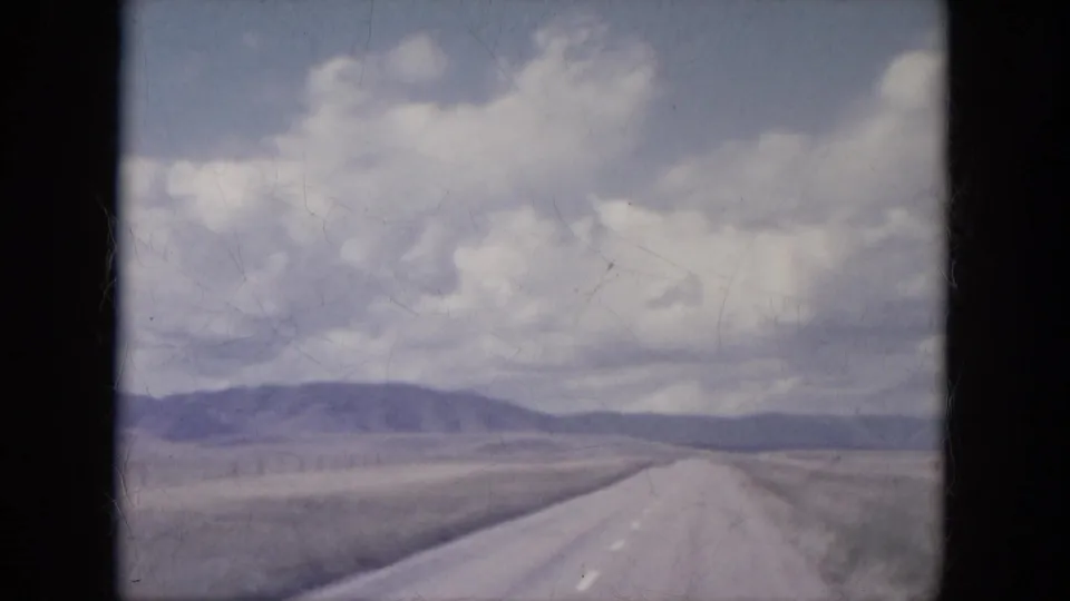 Straight South Dakota highway receding across open fields toward distant hills in a faded 1960s 8mm frame.
